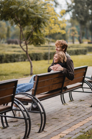 cheerful young man hugging woman and sitting on wooden bench in parkの写真素材