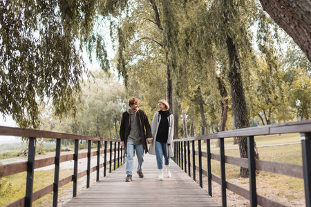 full length of redhead man and blonde woman in coats smiling while holding hands and walking on bridgeの写真素材