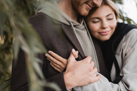 pleased blonde woman hugging with cheerful young man near green leaves on blurred foregroundの写真素材