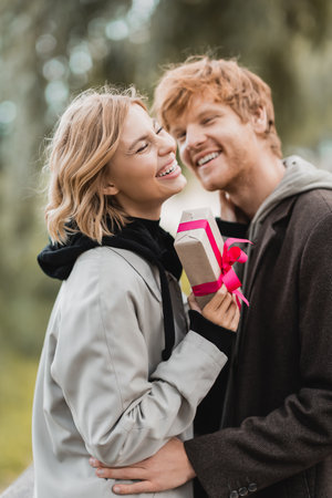 happy woman laughing while holding wrapped gift box near cheerful boyfriend on blurred backgroundの写真素材