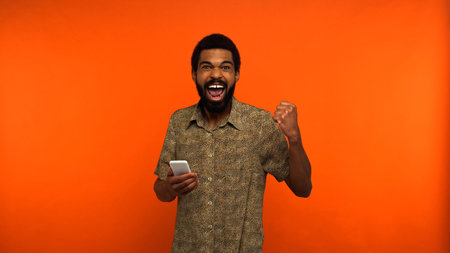 excited african american man with beard holding smartphone on orange background