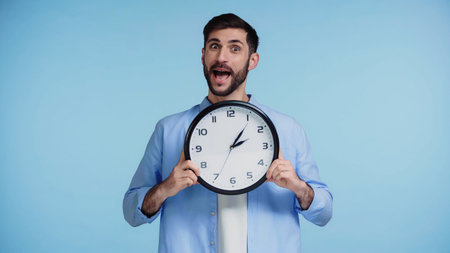 bearded man in shirt holding clock while looking at camera isolated on blue background