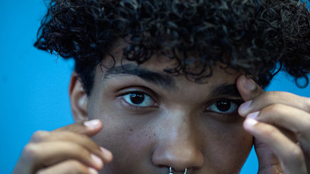 Cropped view of african american man touching eyebrow and looking at camera isolated on blueの写真素材