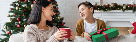 happy woman with cup of warm cocoa looking at cheerful son with christmas present, banner