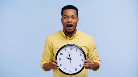 amazed african american man holding clock and looking at camera isolated on blueの写真素材