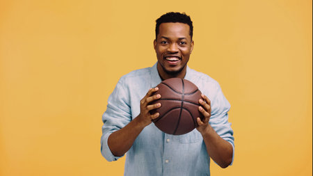 happy african american man holding basketball isolated on yellowの写真素材