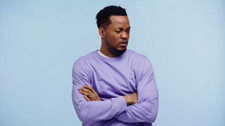 offended african american man in purple pullover standing with crossed arms isolated on blue