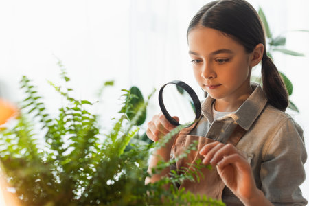 Kid in apron holding magnifying glass near green plant at homeの写真素材