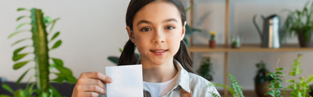 Smiling kid holding sticky note near blurred plants at home, bannerの写真素材