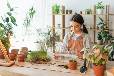 Preteen child in apron holding test tubes near plants and flowerpots at homeの写真素材