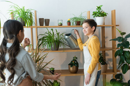 Smiling boy watering plant near blurred friend at homeの写真素材