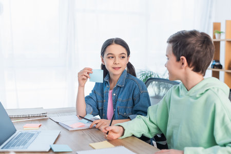 smiling girl holding empty sticky note near blurred boy and laptop on deskの写真素材