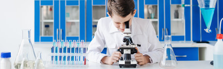 boy in white coat looking into microscope near flasks and test tubes in chemical lab, bannerの写真素材
