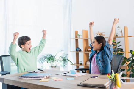 excited kids showing win gesture near notebooks and paper notes on table at homeの写真素材
