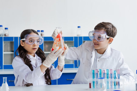 happy preteen kids in white coats looking at camera near microscope and test tubes with flasks