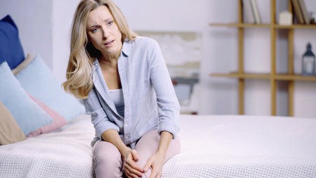 blonde woman suffering from pain in knee while sitting on bed at homeの写真素材