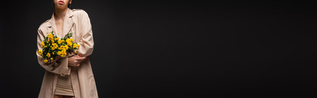 Cropped view of model in beige trench coat holding bouquet of chrysanthemums isolated on black, bannerの写真素材