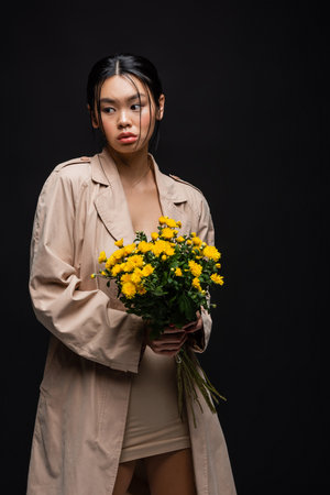 Young asian model in dress and trench coat holding yellow flowers isolated on blackの写真素材