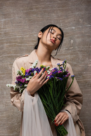 Low angle view of stylish asian woman in trench coat holding wildflowers on abstract brown backgroundの写真素材