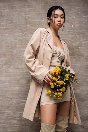 Stylish asian woman in dress and boots holding yellow flowers on abstract brown backgroundの写真素材