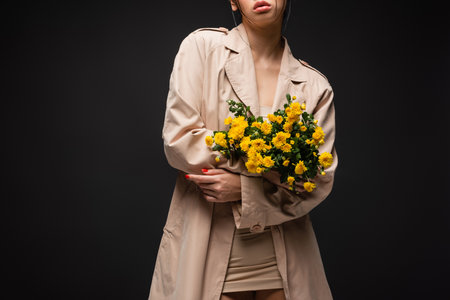 Cropped view of young woman in trench coat holding chrysanthemums isolated on blackの写真素材