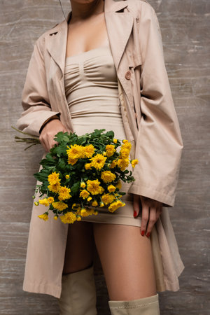 Cropped view of stylish woman in beige dress and trench coat holding yellow flowers on abstract brown backgroundの写真素材