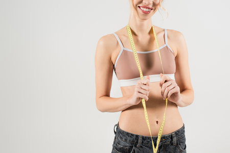 cropped view of happy and fit woman in sports top holding measuring tape isolated on greyの写真素材
