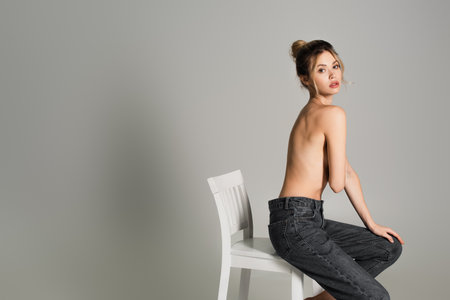 young half nude woman in jeans sitting on white chair and looking at camera isolated on greyの写真素材