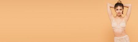 fit woman in lace bra posing with hands behind head and smiling at camera isolated on beige, bannerの写真素材