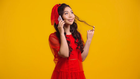 happy asian woman in red dress talking on smartphone while twirling hair isolated on yellowの写真素材