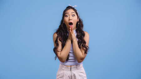 shocked and young asian woman with opened mouth and praying hands isolated on blue
