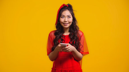 happy asian woman in red dress texting on smartphone isolated on yellowの写真素材