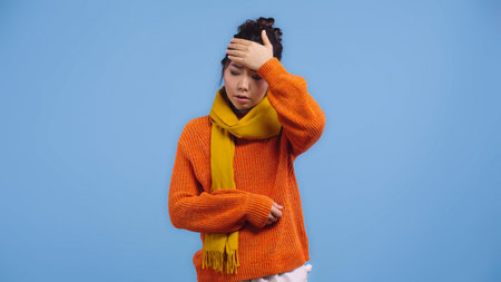 sick asian woman in orange sweater and scarf touching forehead isolated on blueの写真素材