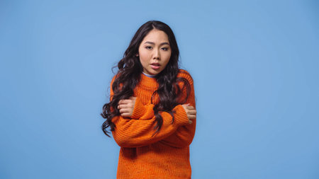 brunette and young asian woman in sweater feeling cold isolated on blue