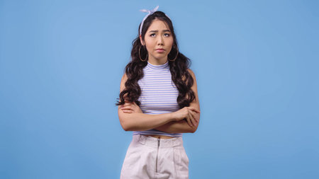frustrated asian woman in headband with bow standing with crossed arms isolated on blue