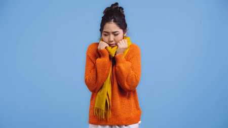 sick asian woman in orange sweater and scarf feeling cold isolated on blueの写真素材