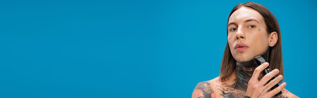 young and pierced man with tattoos shaving with electric razor isolated on blue, bannerの写真素材