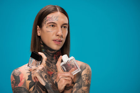 shirtless and tattooed man holding bottles with perfume isolated on blue