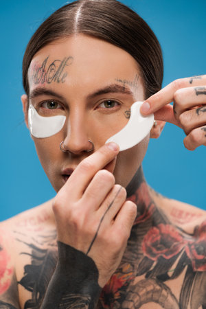tattooed young man applying eye patches and looking at camera isolated on blueの写真素材