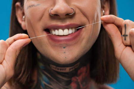 cropped view of cheerful and tattooed man flossing white teeth isolated on blueの写真素材