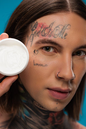 close up of tattooed young man holding container with cosmetic cream isolated on blue