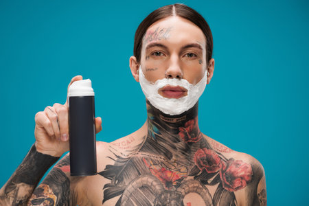 shirtless and tattooed young man with shaving foam on face holding can isolated on blueの写真素材