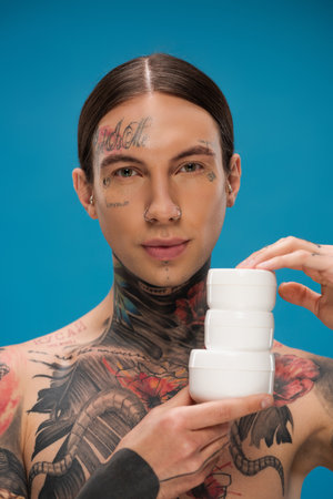 tattooed young man holding containers with cosmetic cream isolated on blueの写真素材
