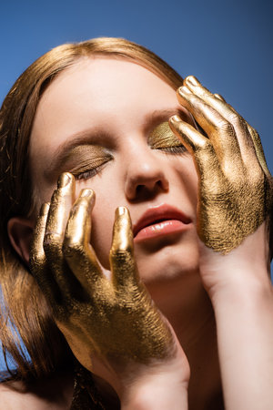 Portrait of young woman with makeup and golden paint on hands touching face isolated on blueの写真素材