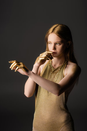 Pretty woman in golden paint and dress touching lips and posing isolated on greyの写真素材
