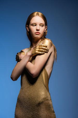 Fair haired model in dress and golden paint on hands touching shoulders isolated on blueの写真素材