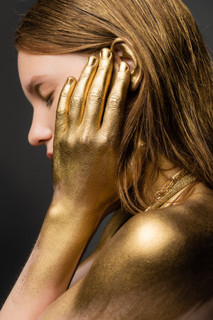 Side view of young woman in golden paint touching ear isolated on greyの写真素材