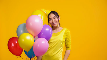 happy asian woman in party cap holding colorful balloons isolated on yellowの写真素材