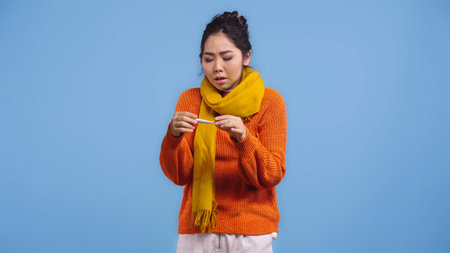 sick asian woman in sweater and scarf holding digital thermometer isolated on blueの写真素材