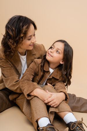 joyful woman and child in stylish suede jackets looking at each other on beige backgroundの写真素材